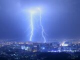 Breathtaking thunderstorm with lightning bolts over modern illuminated city at night with purple sky