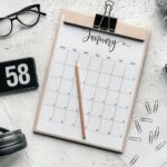 Top view composition of clipboard with calendar and pencil placed on desk amidst stationery and eyeglasses