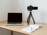 Modern workplace consisting of netbook camera on tripod near opened planner with pen on wooden table