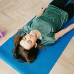 From above of young slender lady in casual clothes and wristwatch resting with closed eyes on blue yoga mat near dumbbells on floor in flat