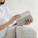 Crop unrecognizable female in white shirt sitting on sofa and opening notepad in light room