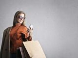 Smiling female in trendy wear and stylish sunglasses standing with paper shopping bags on background of gray wall and browsing mobile phone