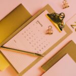 From above stylish workplace consisting of clipboard with calendar and golden notebook on pink background