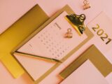 From above stylish workplace consisting of clipboard with calendar and golden notebook on pink background