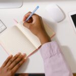 From above of crop unrecognizable African American employee writing in empty organizer at white table