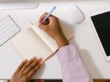 From above of crop unrecognizable African American employee writing in empty organizer at white table