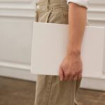 Side view of unrecognizable female in stylish jeans with white blank notebook in hand standing in light room near door