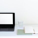 Laptop with blank screen and glass of water arranged on table near white daily planner and pencil in modern workspace