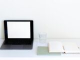 Laptop with blank screen and glass of water arranged on table near white daily planner and pencil in modern workspace