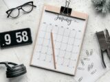 Top view composition of clipboard with calendar and pencil placed on desk amidst stationery and eyeglasses