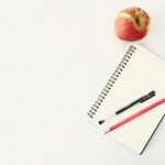 Top view composition of pen and pencil placed on spiral opened notebook with blank sheets near red ripe apple on white desk