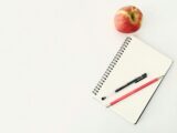 Top view composition of pen and pencil placed on spiral opened notebook with blank sheets near red ripe apple on white desk