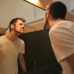 Low angle side view of young bearded male in casual shirt standing in bathroom and looking at with frown mirror in morning