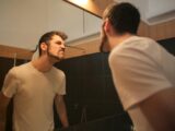 Low angle side view of young bearded male in casual shirt standing in bathroom and looking at with frown mirror in morning