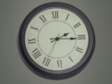 Round black mechanical clock with Roman numerals hanging on gray wall