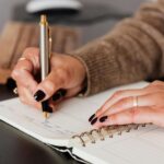 Crop unrecognizable female with stylish manicure sitting at black desk with keyboard and smartphone and taking notes with silver pen in notepad