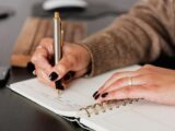 Crop unrecognizable female with stylish manicure sitting at black desk with keyboard and smartphone and taking notes with silver pen in notepad