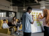 Photo of a Man and a Woman Writing on a Whiteboard with Sticky Notes