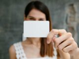 Young blurred female showing white blank business card and looking at camera in light room