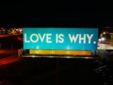 Blue billboard saying Love is why placed on road surrounded by cars and street lights against black night